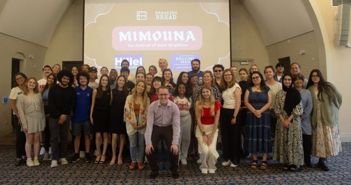 A large, diverse group of Jewish and Muslim college students standing together at a Rollins College Mimouna celebration. They are positioned in front of a projection screen that reads "Mimouna: The Festival of Good Neighbors" with Hillel and Muslim Student Union logos.